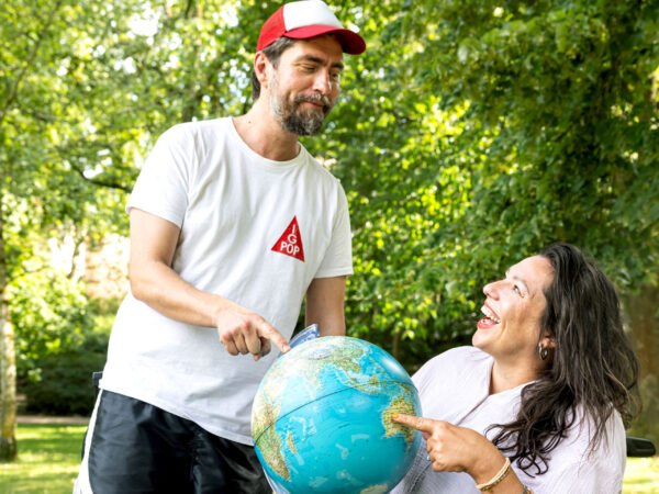 David reicht Kim im Rollstuhl lachend einen Globus, im Hintergrund Bäume.