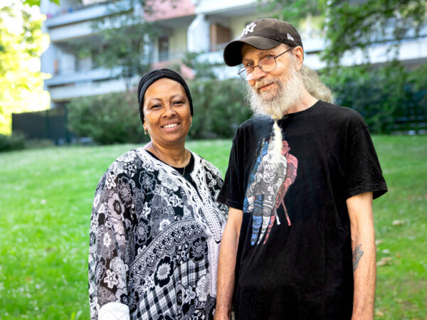 Sandra und Klaus stehen lächelnd nebeneinander auf einer Wiese vor einem Mehrfamilienhaus.