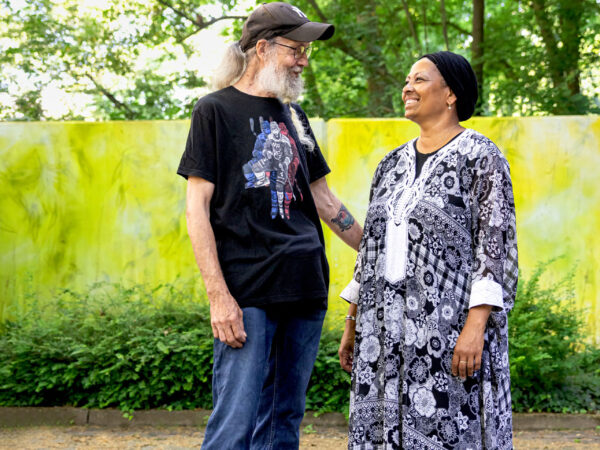 Sandra und Klaus auf einem gepflasterten Weg vor einer gelben Mauer zwischen Bäumen.