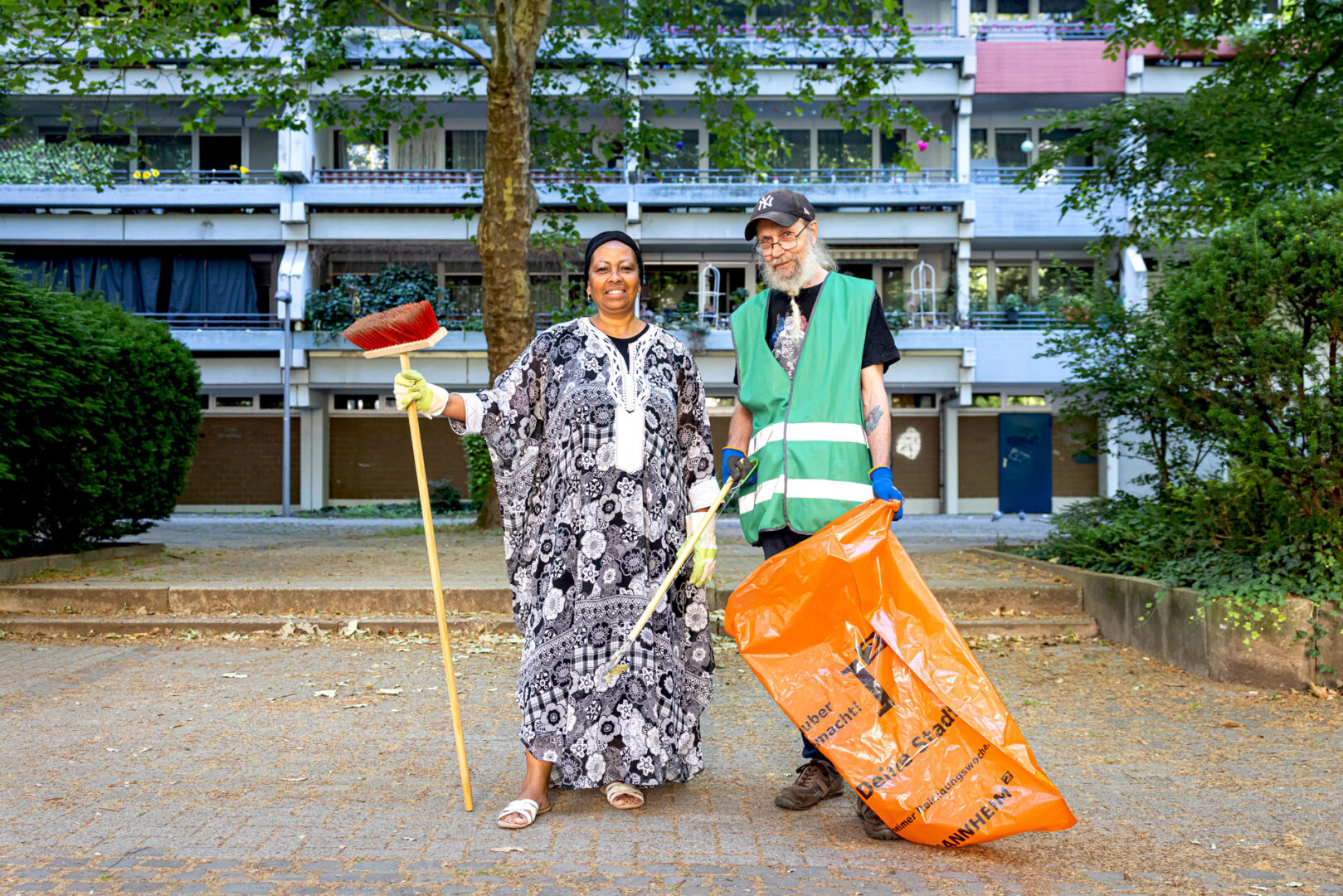 Sandra mit Besen und Klaus mit Warnweste und orangefarbenem Müllsack vor einem Wohngebäude.