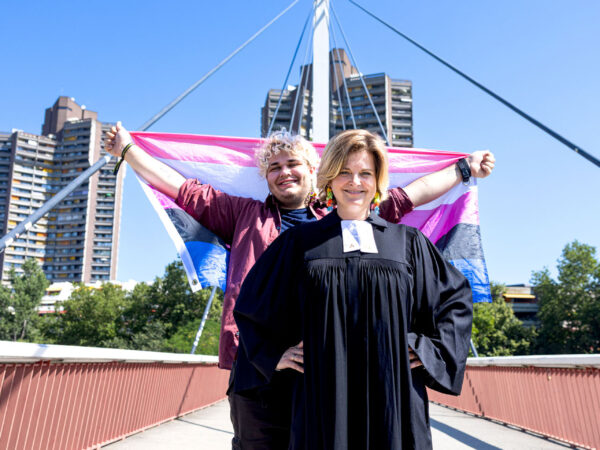 Nina im schwarzen Talar steht auf einer Brücke, hinter ihr hält Panda eine Trans-Flagge in die Höhe.
